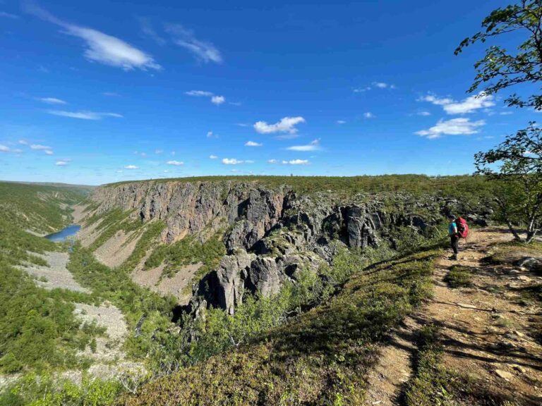 Jylhä tunturimaisema, kuvan oikeassa reunassa vaeltaja rinkka selässään. Sininen taivas, jolla untuvaisia valkoisia pilviä.